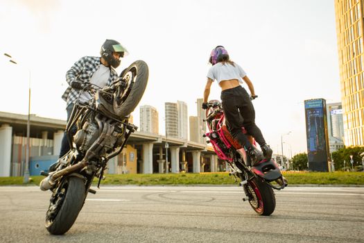 Two riders performing motorcycle stunts in a cityscape setting.