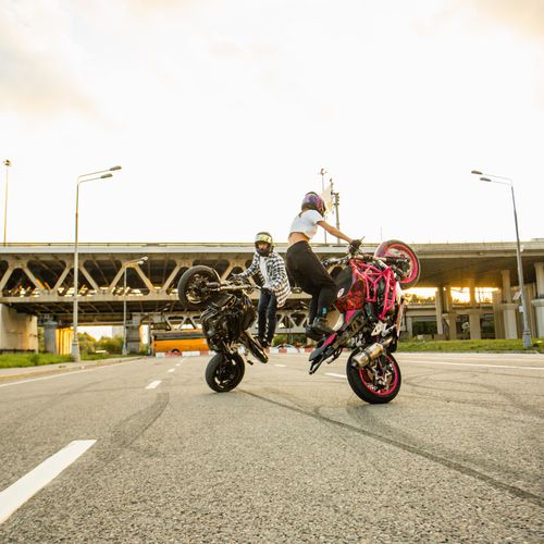 Free Two motorcyclists performing stunts on city streets under a bridge at sunset. Stock Photo