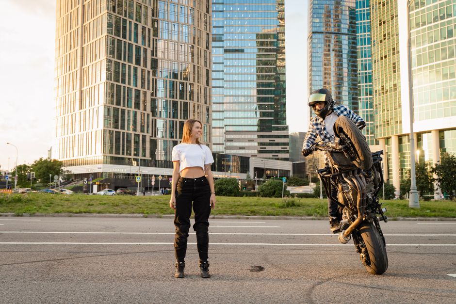 Motorcyclist doing a wheelie in front of skyscrapers while a woman watches during daylight.