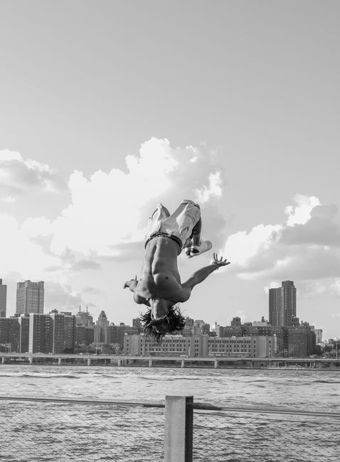 Shirtless man executing a backflip by the waterfront with New York City skyline in the background.