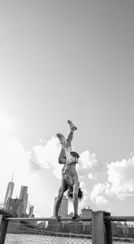 Black and white photo of a shirtless man doing a handstand on a fence with New York City skyline in the background.