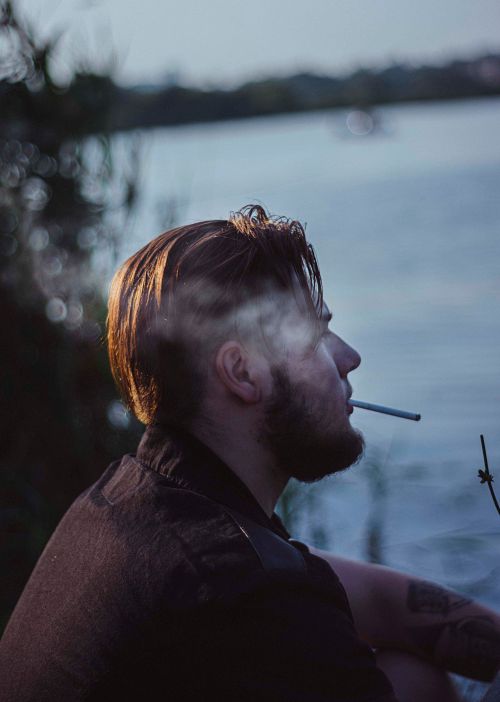 Free A man smokes by the serene lake, captured in a thoughtful side view. Stock Photo