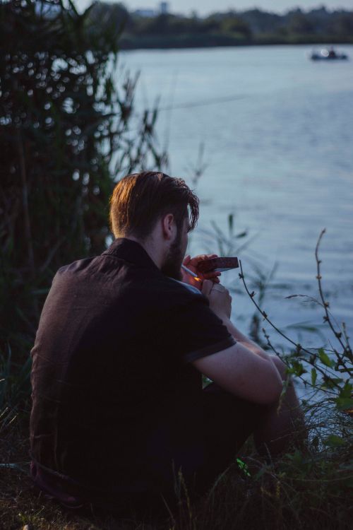 Free A man sits by a riverside, enjoying a quiet moment while smoking in nature. Stock Photo