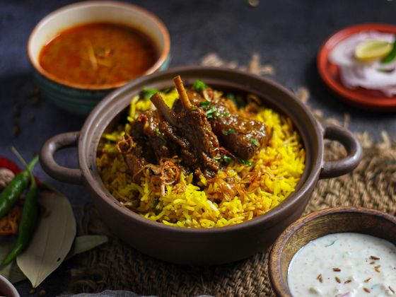 Clay pot of saffron mutton biryani with bone-in meat and fried onions, raita and curry bowl