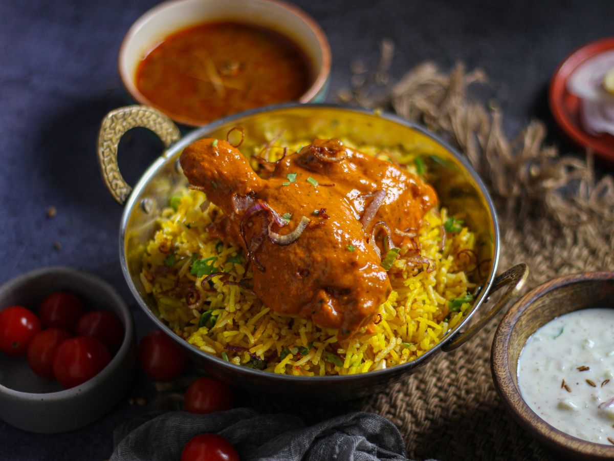 Masala-coated chicken leg on yellow rice in a brass pot with raita and gravy