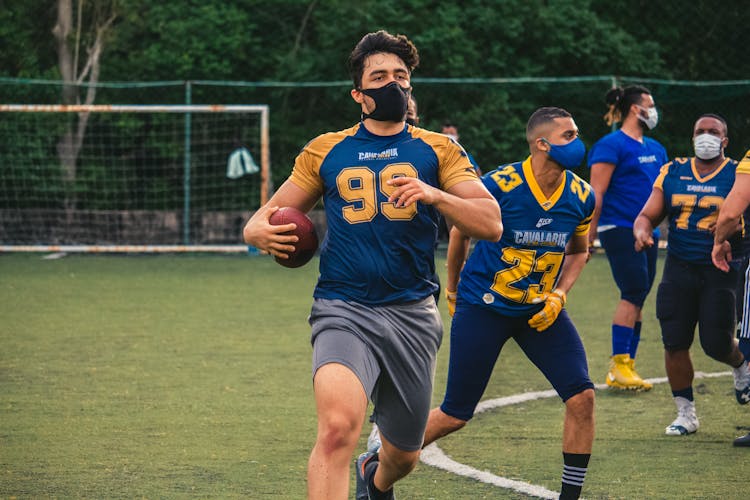 A Man Wearing A Face Mask Running With A Rugby Ball