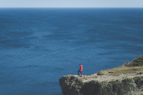 Man in Red Jacket Standing on Cliff Edge Near Body of Water