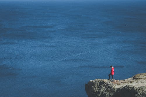 Man in Red Jacket Standing on Cliff Edge Near Body of Water