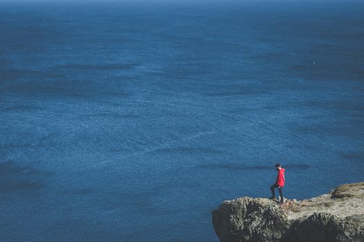 A man in a red jacket stands on a cliff edge overlooking a vast ocean.