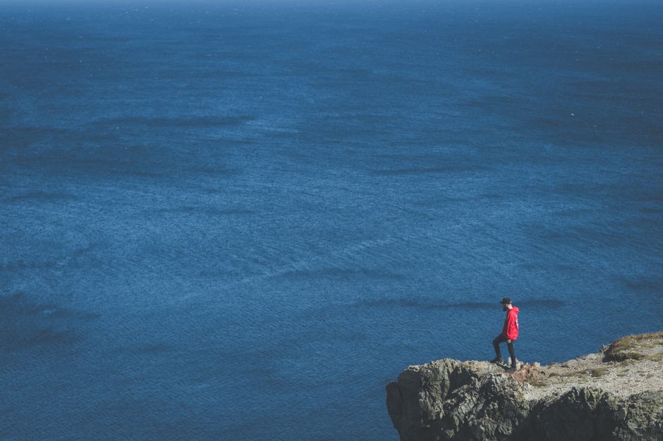 A man in a red jacket stands on a cliff edge overlooking a vast ocean.