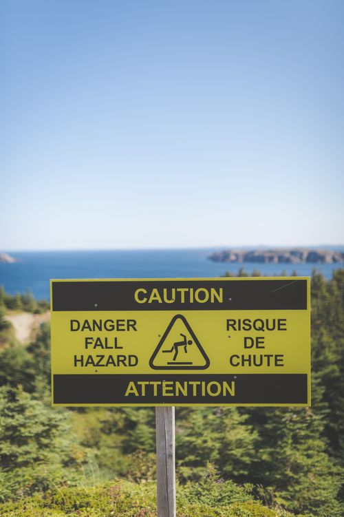 Free Yellow caution sign warns of fall hazard near a scenic ocean view with green foliage. Stock Photo