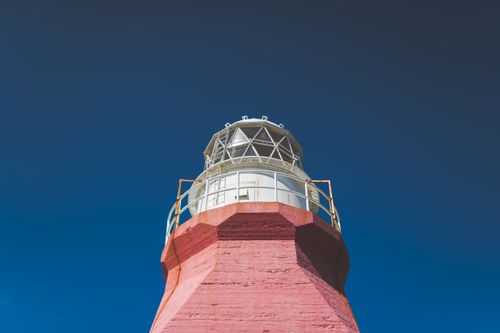 Free A majestic red brick lighthouse captured from a low angle against a clear blue sky, highlighting its architectural beauty. Stock Photo