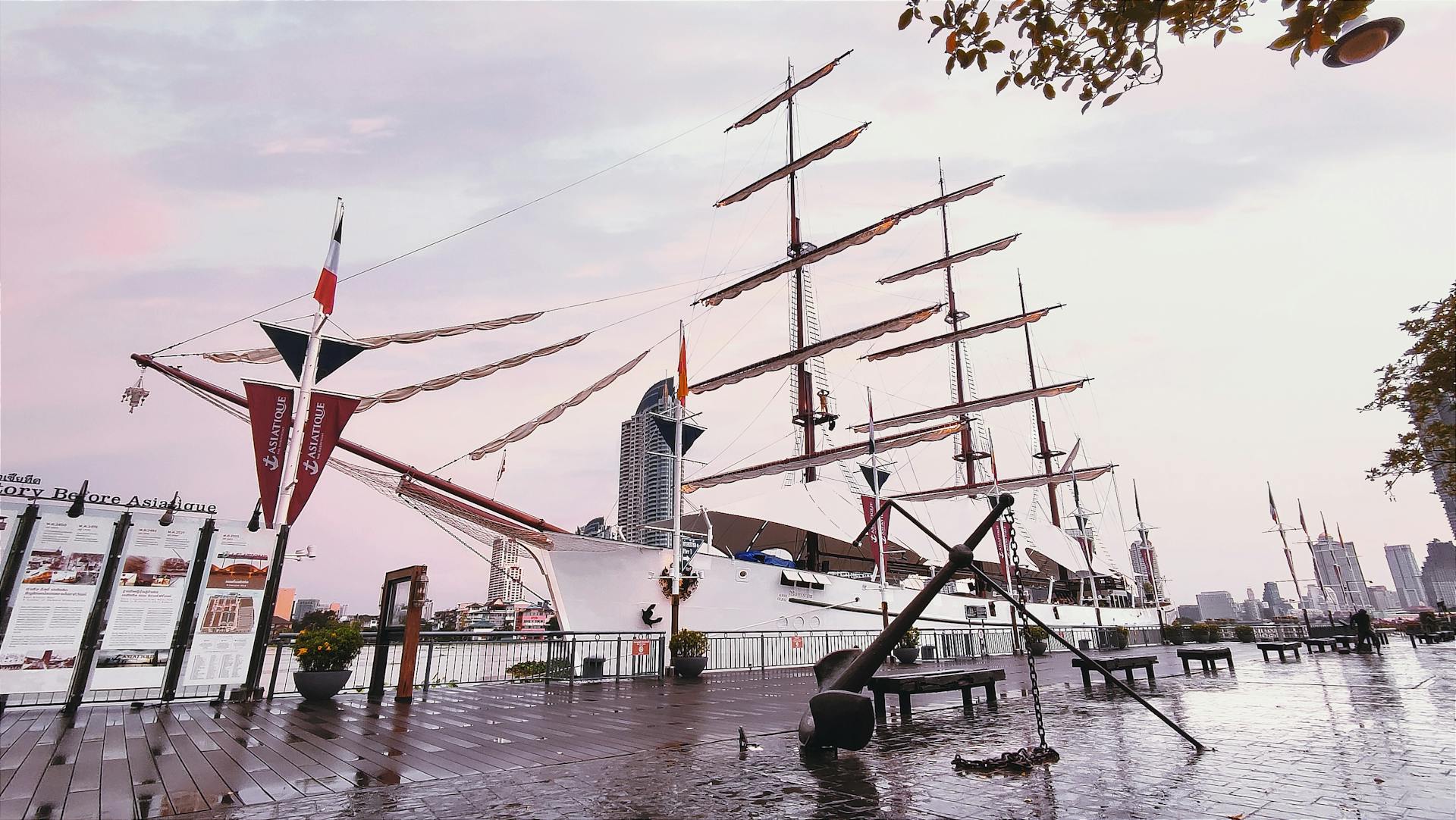 Tall ship docked in Bangkok's riverside port during dusk with a tranquil skyline.