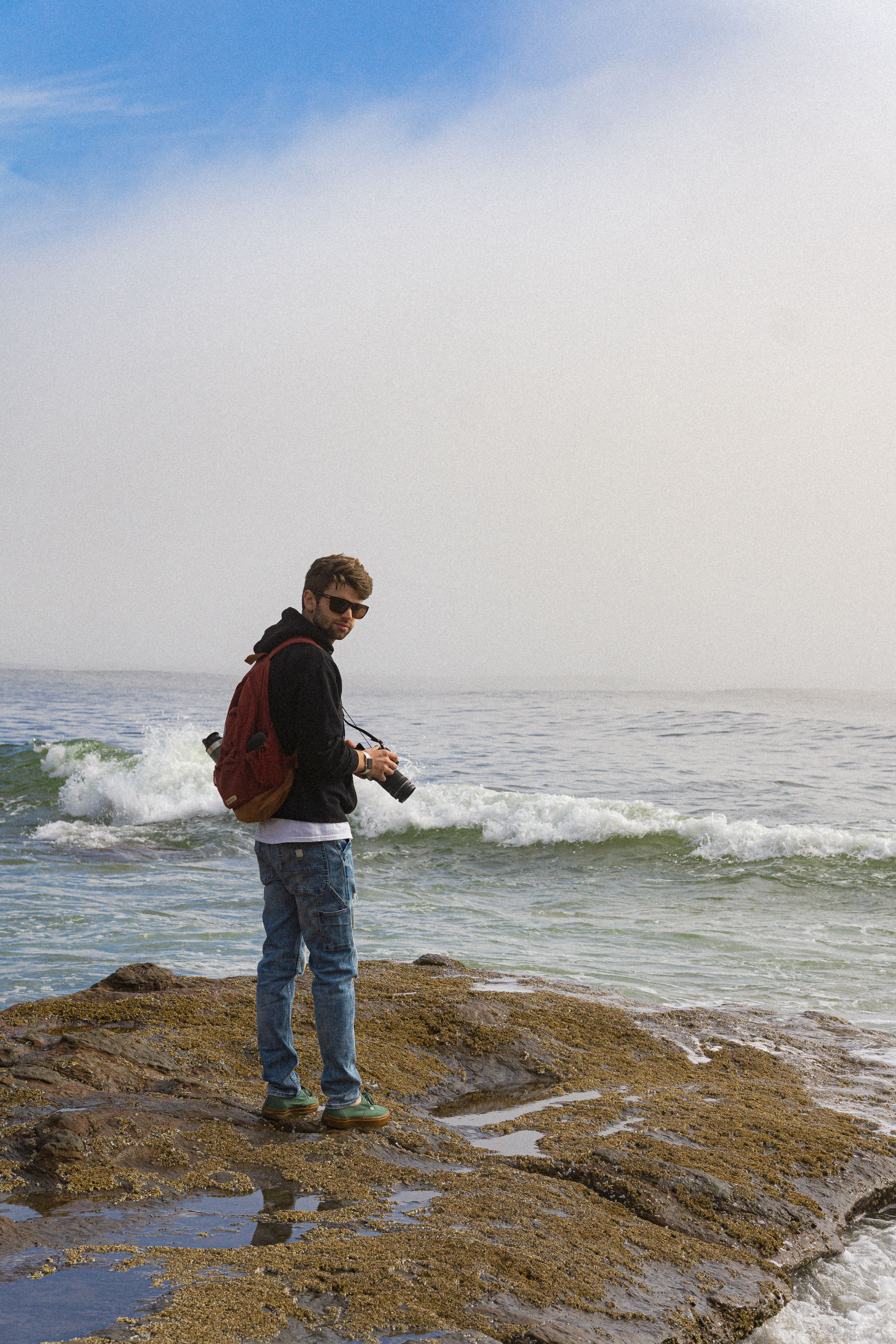 Man with camera and backpack standing on rocky coast capturing sea waves.