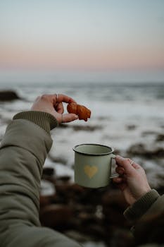 Cozy morning by the sea holding a mug and pastry at sunrise for a peaceful start.