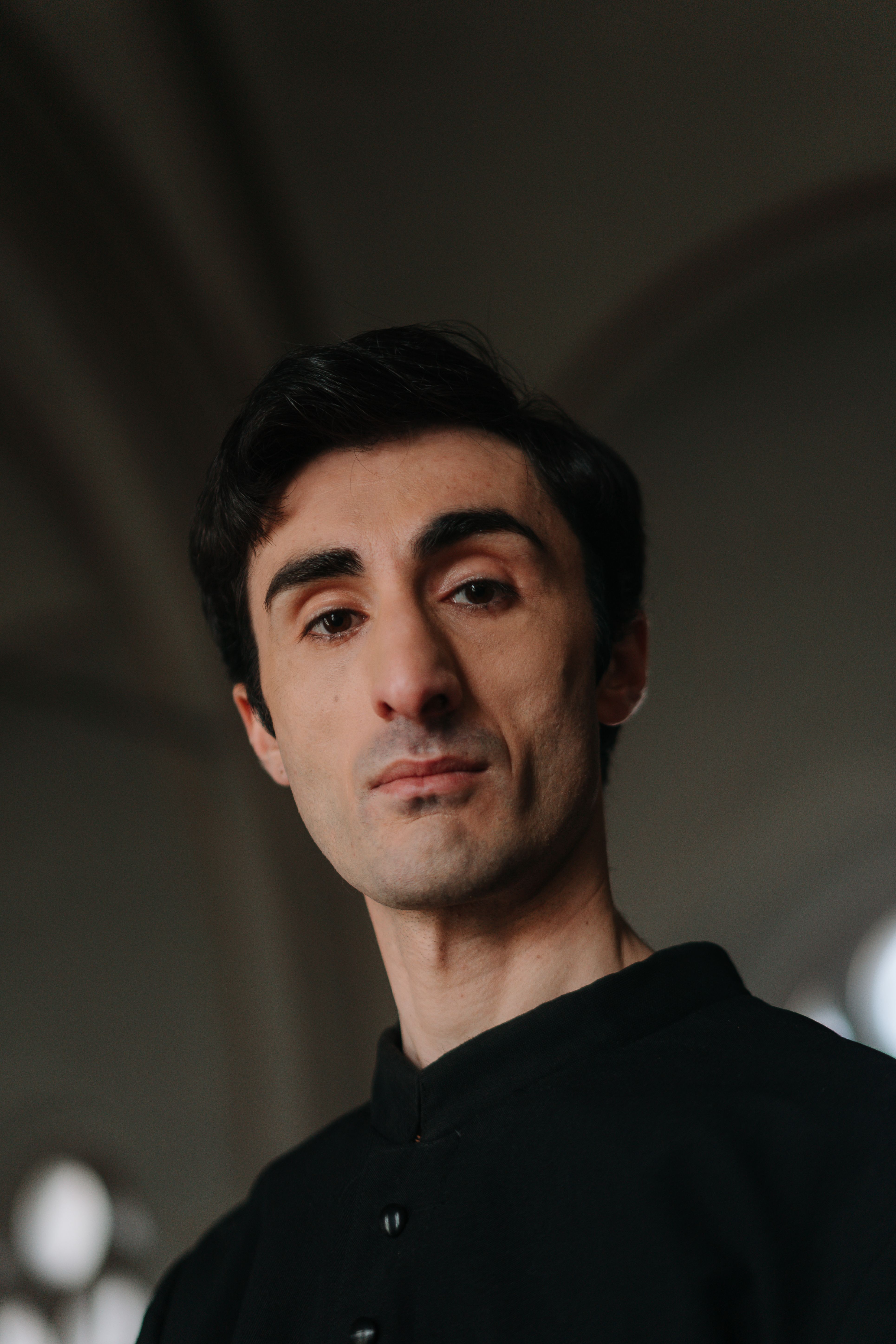 Elegant portrait of a man in black clothing, thoughtfully gazing indoors with soft lighting.
