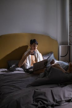 Shirtless man enjoys morning coffee in bed while reading the newspaper.