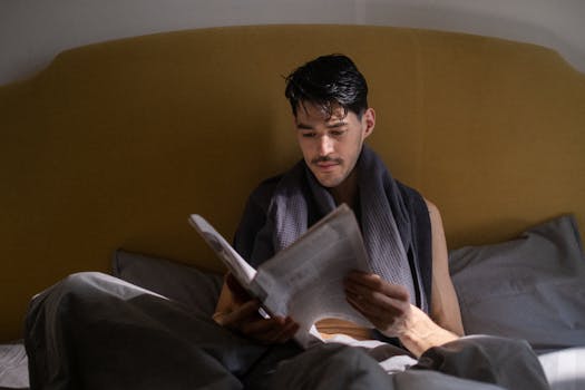 A man with dark hair reads a newspaper in bed during a quiet morning routine.