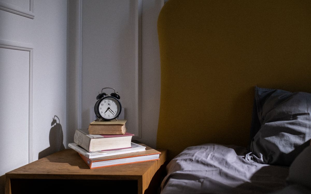 A cozy bedroom setting with books and an alarm clock on a nightstand next to a bed.