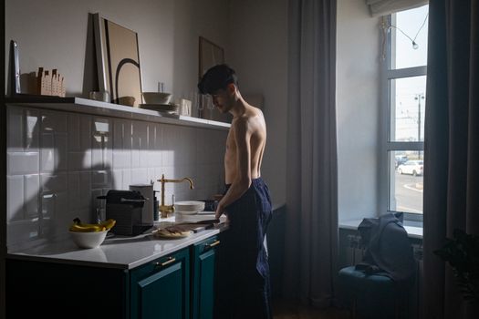 A shirtless man prepares breakfast in a sunlit kitchen during an early morning routine.