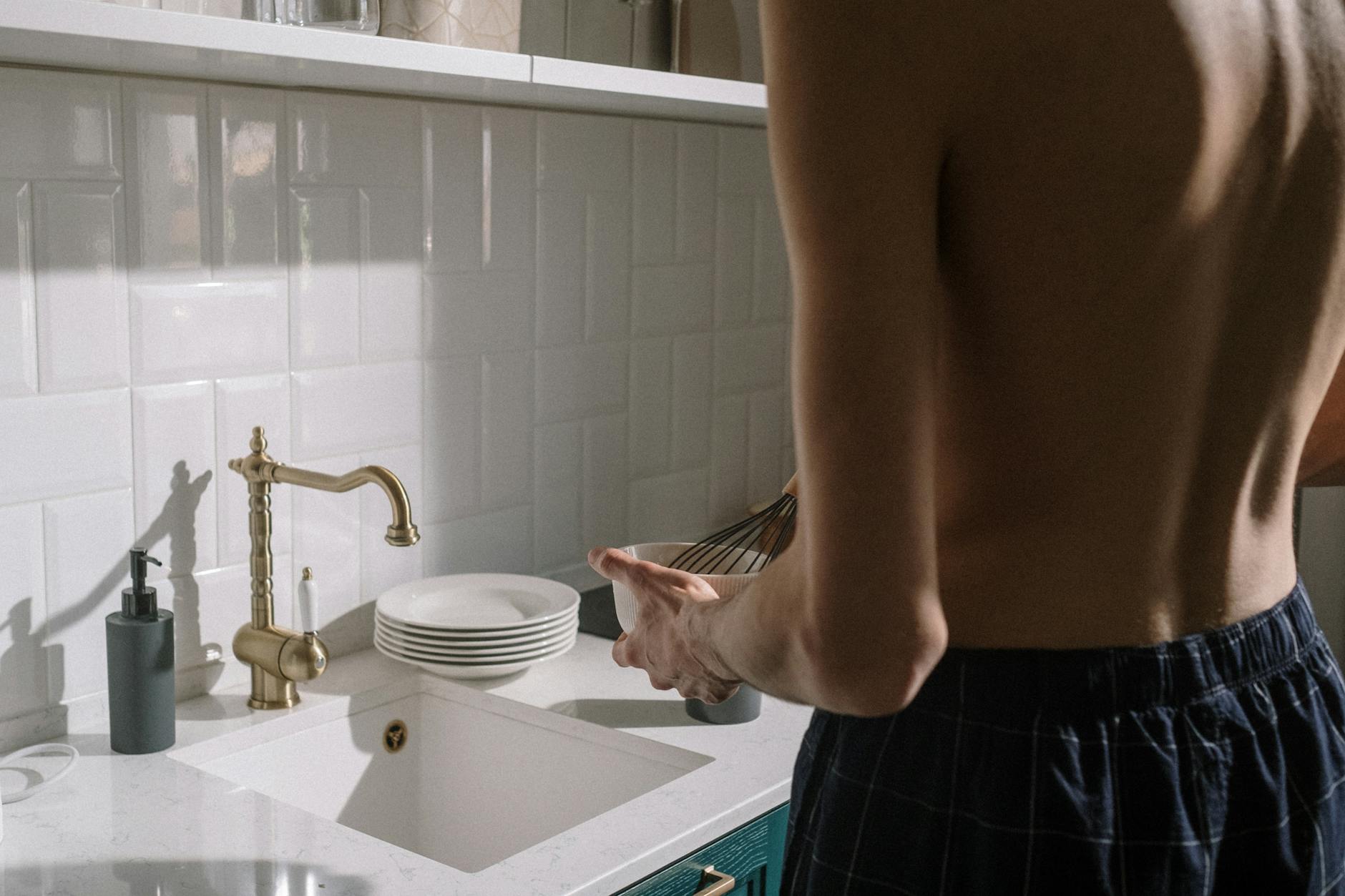 A young man prepares breakfast in a minimalist kitchen, whisk in hand.