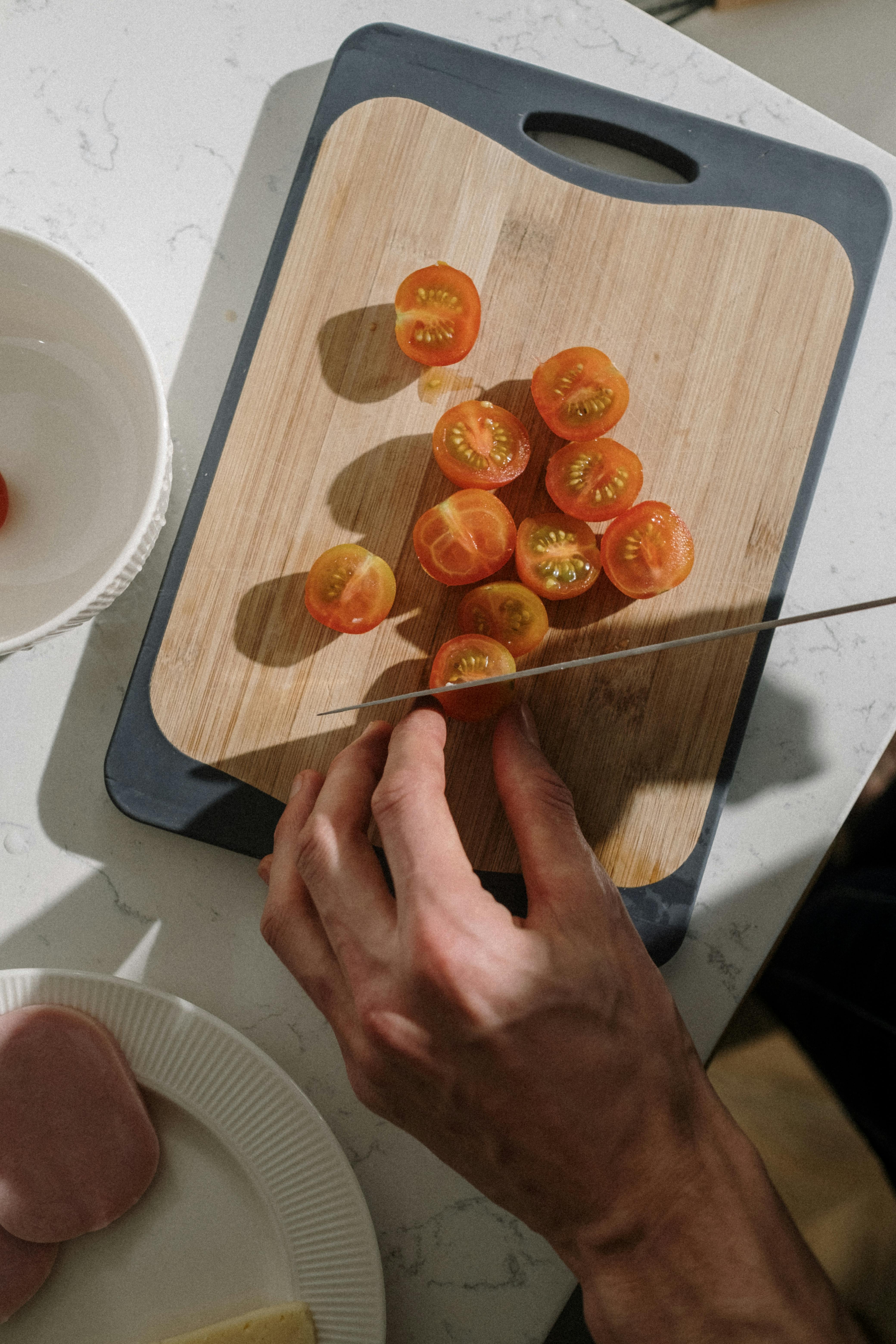 A hand slicing fresh cherry tomatoes on a wooden cutting board with a knife.