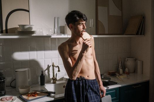 Shirtless young man bites toast in bright kitchen morning scene.