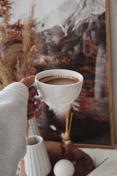 White cup of coffee held in a cozy indoor setting with rustic decor and autumnal tones.