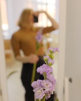 Creative mirror selfie with a focus on lavender flowers and blurred reflection of a woman indoors.