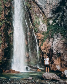 A man stands by a picturesque waterfall, surrounded by rocks and lush greenery, capturing an adventurous moment.