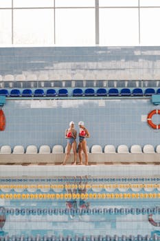 Two female swimmers in blue swimwear by an indoor pool, preparing for training.