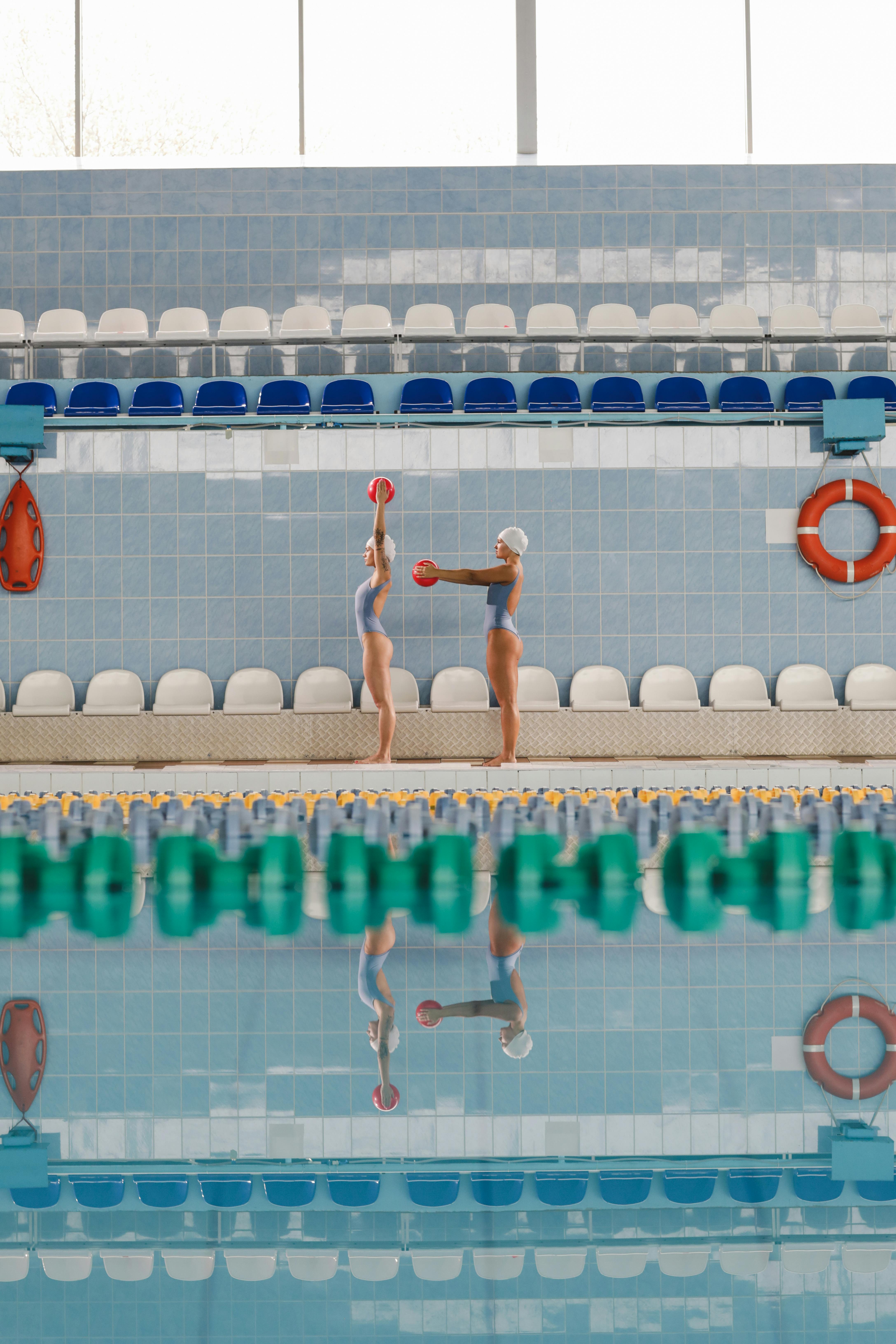 Two women in swim caps practice synchronized movements by the pool reflecting in the water.