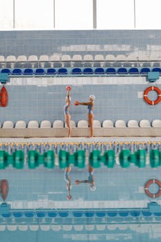 Two women in swim caps practice synchronized movements by the pool reflecting in the water.