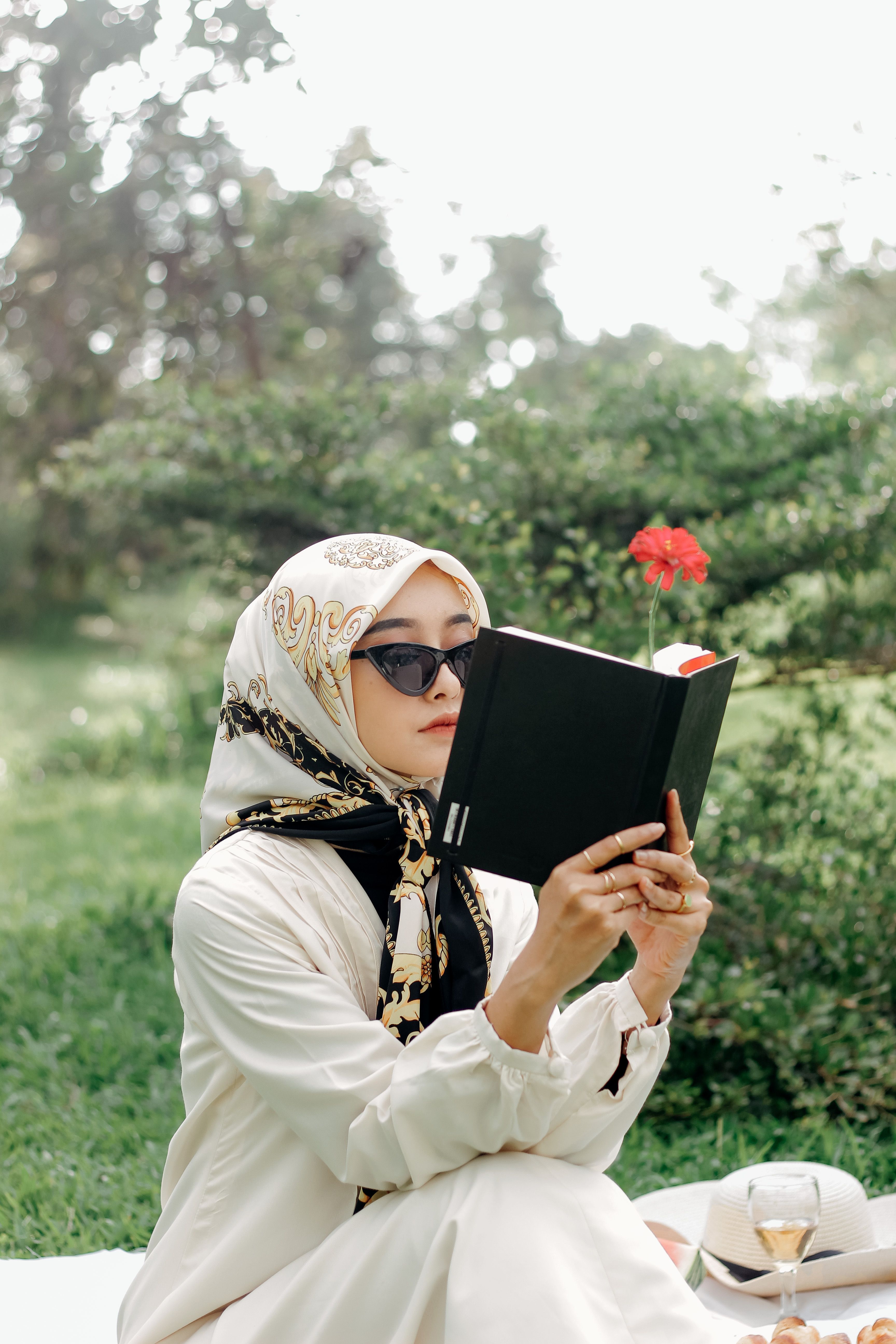 Woman wearing a headscarf reading a book outdoors in a sunny park.