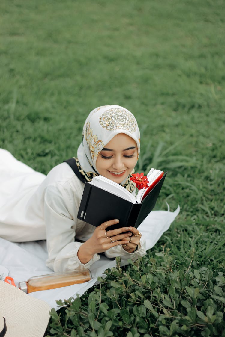 A Woman Wearing White Hijab Lying On Front On Grass Reading A Book