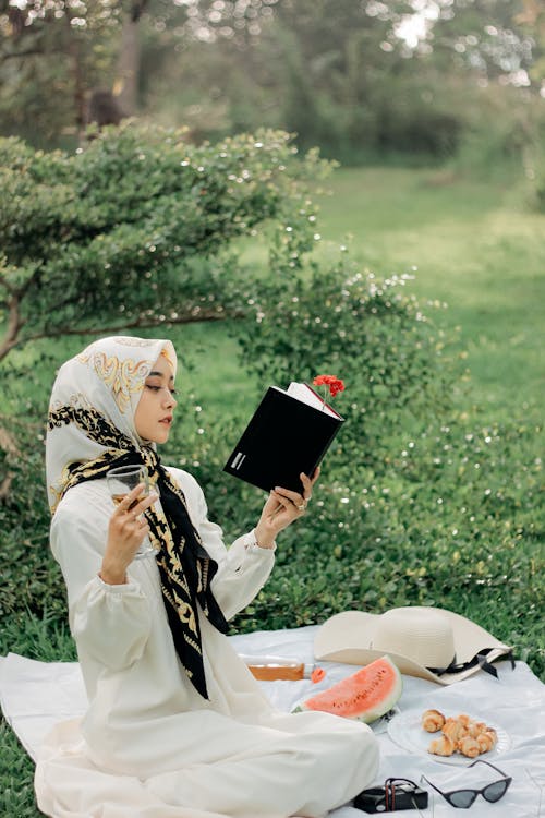 A Woman Wearing White Hijab Sitting on Picnic Blanket Reading a Book