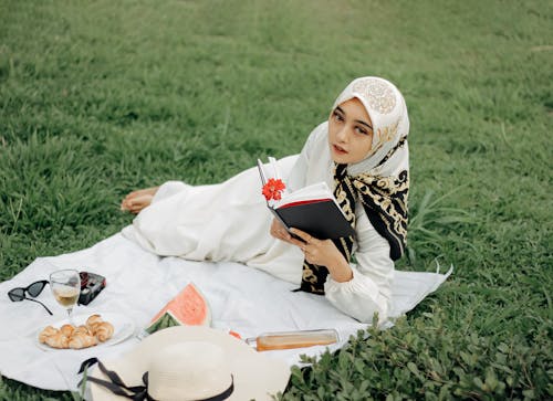 A Woman Wearing White Hijab Lying on Side on Picnic Blanket Reading a Book
