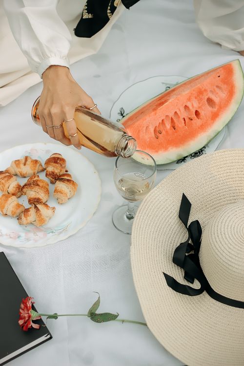 Free Stylish picnic setup with croissants, watermelon, and rosé on a white blanket outdoors. Stock Photo