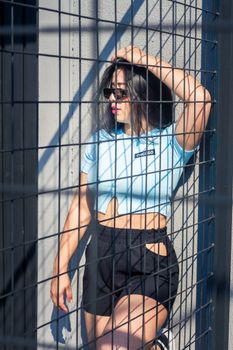 Chic young woman with sunglasses standing beside a metal fence in urban setting.
