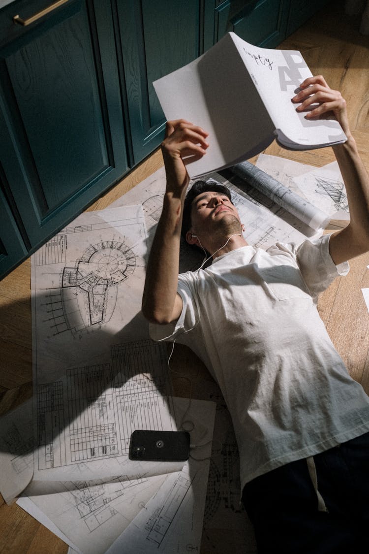 Adult Man Laying Down On Floor Among Drawings And Reading Book