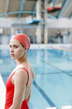 Woman in red swimwear and cap standing by an indoor swimming pool.