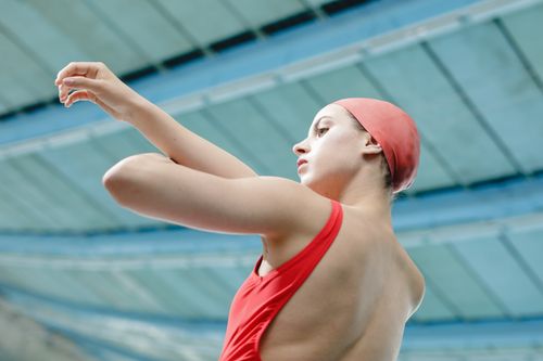 Free Caucasian female swimmer in red swimsuit and cap, prepping indoors. Stock Photo
