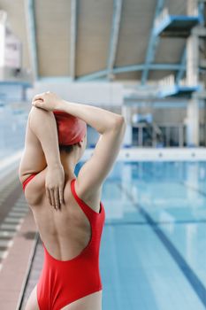 Woman in red swimwear and cap stretching by a swimming pool indoors.
