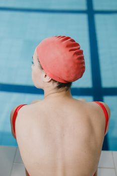 A woman in swimwear and cap sits by the pool edge, back view.