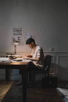 Man studying intensely at a desk, surrounded by drawings and illuminated by a desk lamp.
