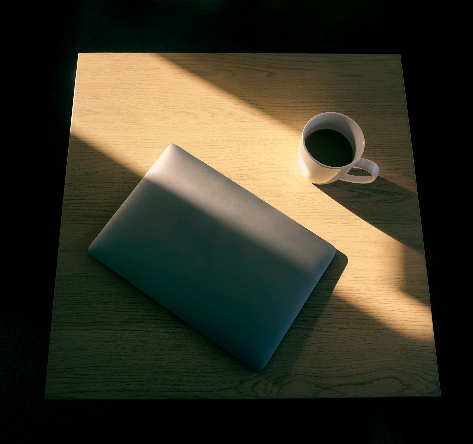 Late afternoon light across a wooden table with a closed laptop and a cup of coffee — photo by Kulbir on Pexels