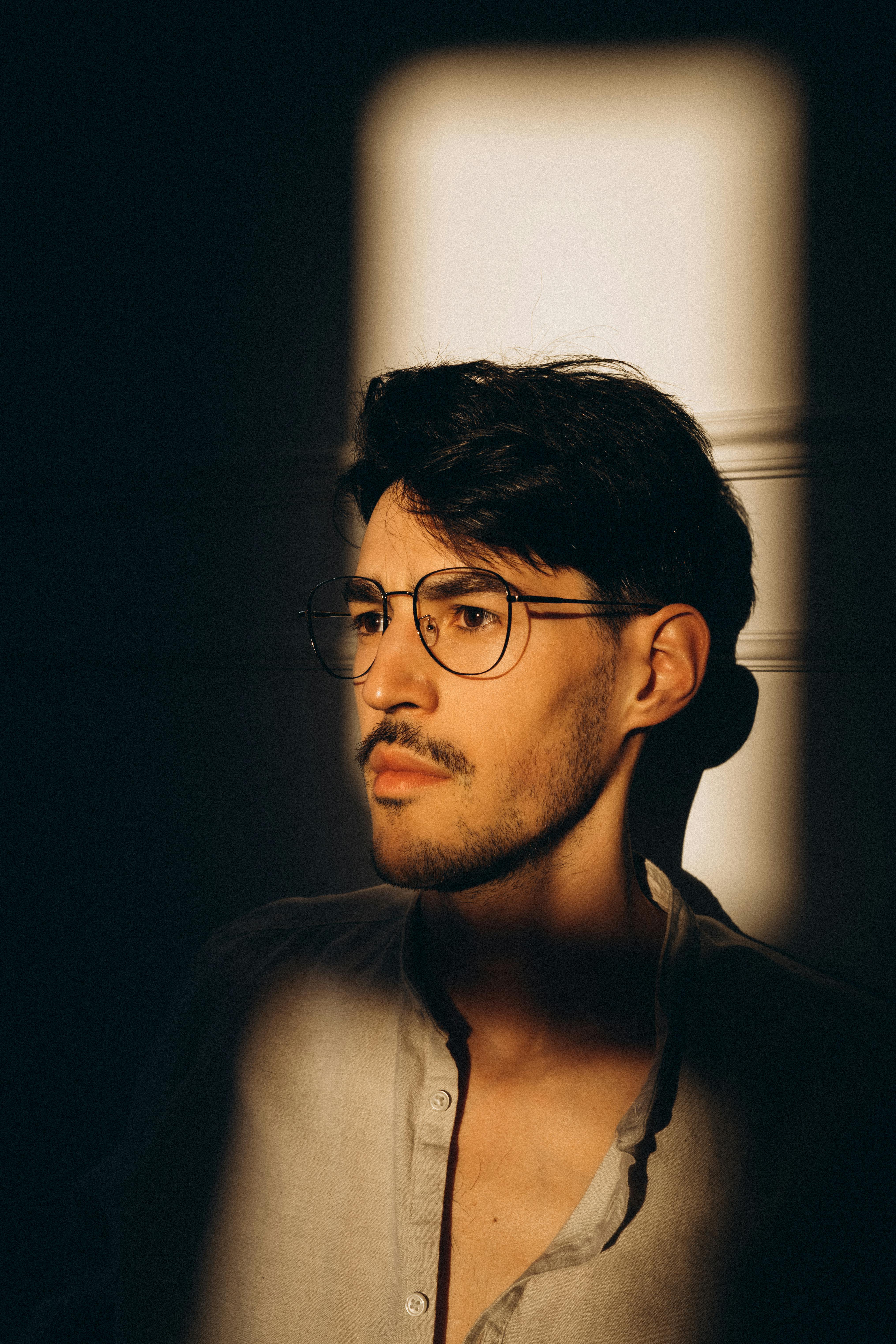 A thoughtful man with glasses captured in dramatic natural light creating a moody, introspective atmosphere.