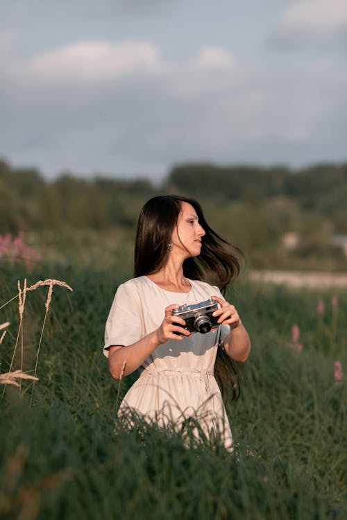 Woman in Beige Dress Holding Camera Standing on Green Tall Grass Field