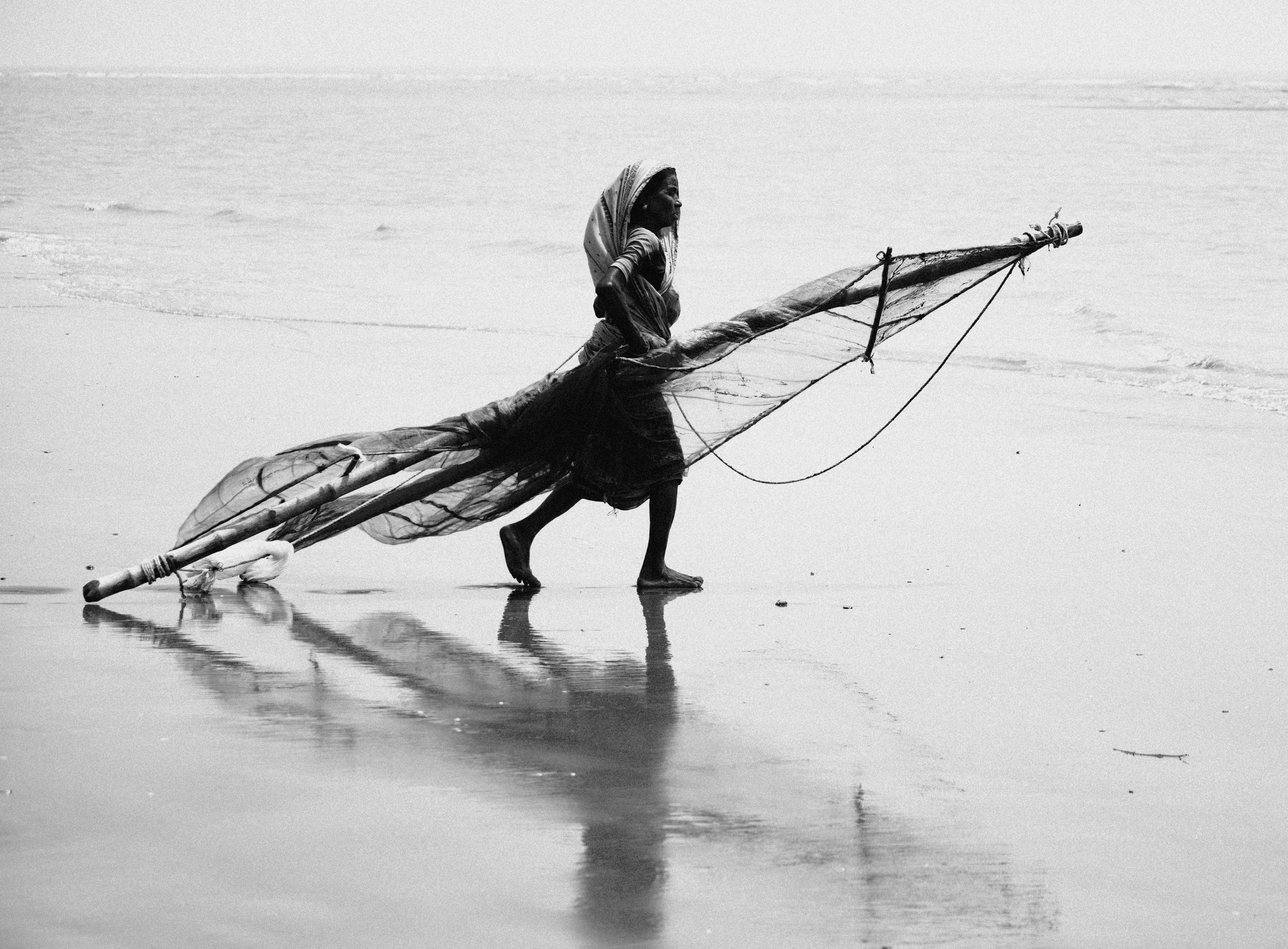 Black and white photo of an African woman gracefully walking with a fishing net on the beach.