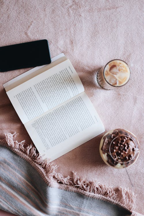 An Open Book and Cellphone with Glasses of Dessert on a Pink Blanket